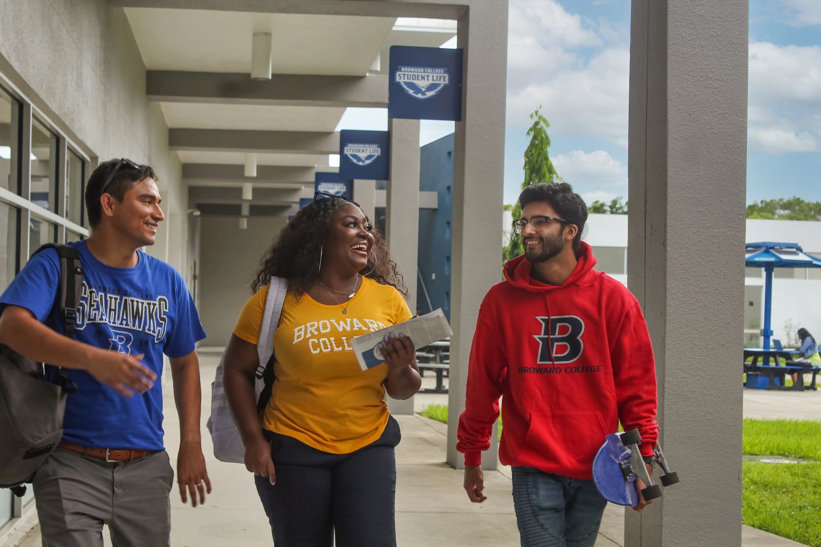 Três estudantes com camisetas do Broward College e dos Seahawks caminham sob a passarela coberta do Student Life