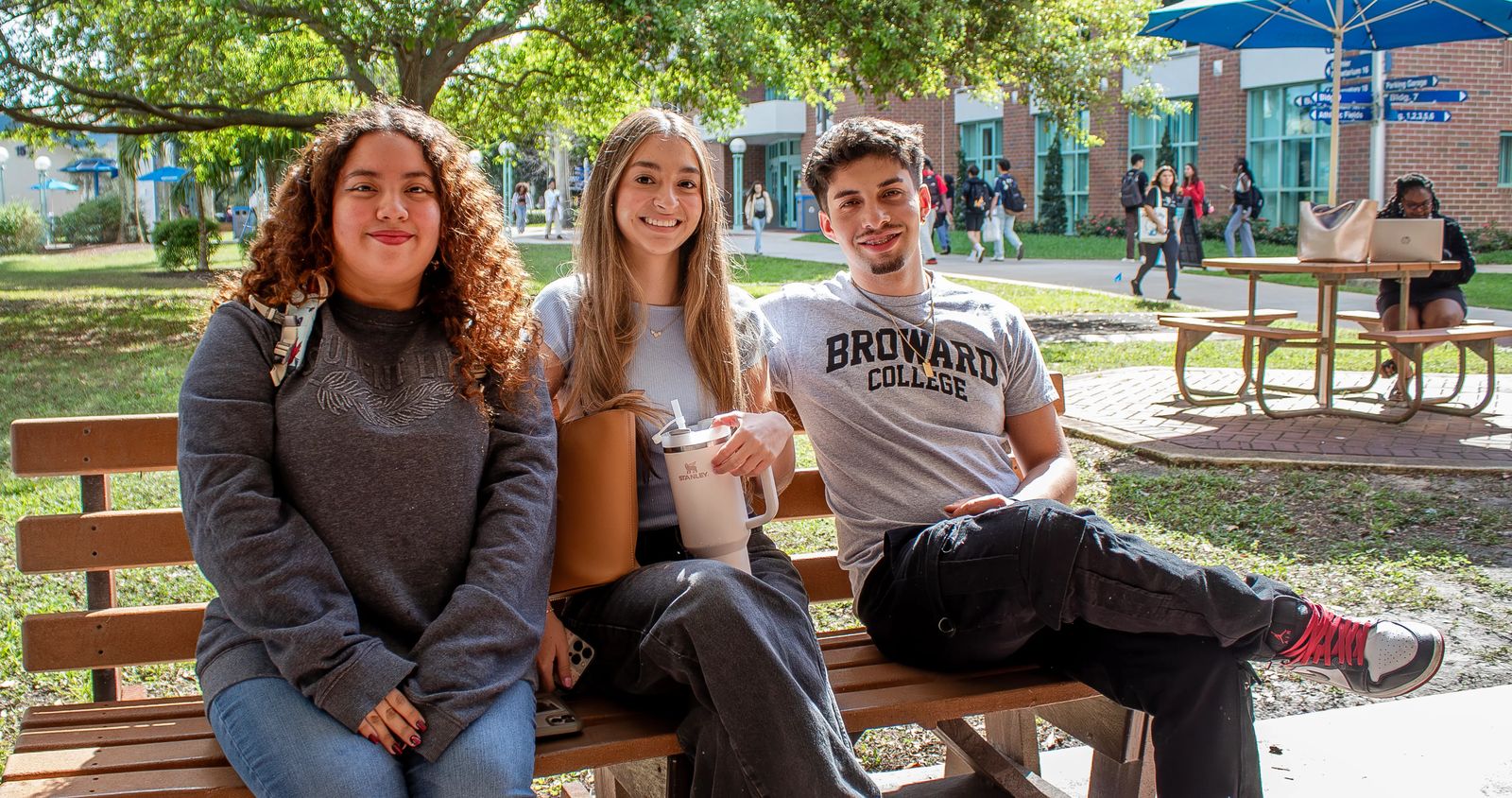 Três estudantes sentados em um banco à sombra das árvores do Central Campus, com prédios de tijolo e colegas ao fundo