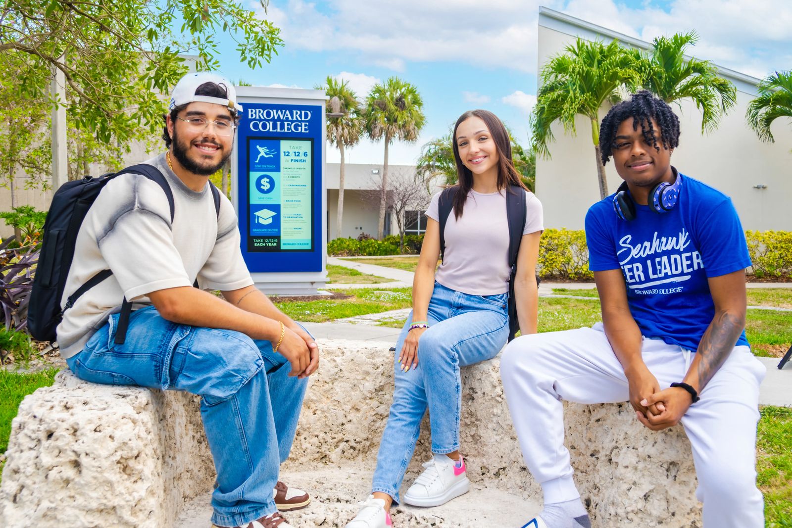 Três estudantes sentados em um muro de pedra coral diante do letreiro do Broward College no South Campus, com palmeiras e céu azul ao fundo