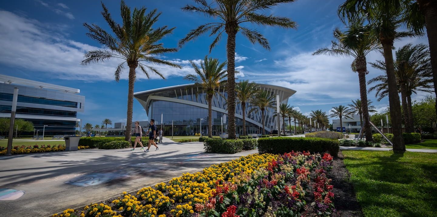The Mori Hosseini Student Union at Embry-Riddle Daytona Beach, with palm trees, flower beds and students walking under a blue sky