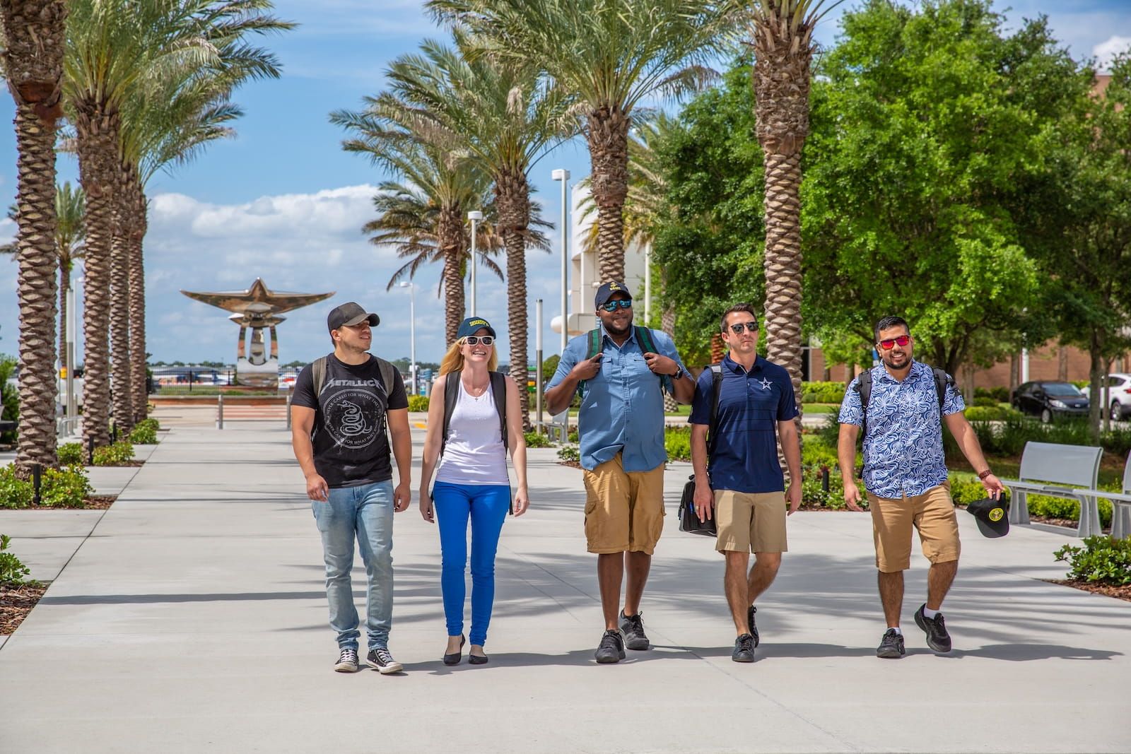 Five student veterans walk along the Legacy Walk at Embry-Riddle Daytona Beach, between palm trees, with the stainless-steel sculpture behind