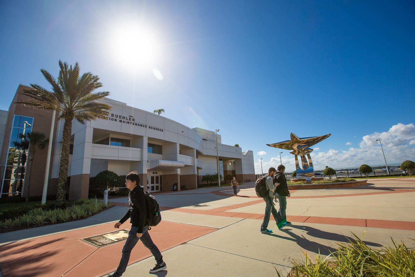 The Emil Buehler Aviation Maintenance Science building on the Legacy Walk at Embry-Riddle Daytona Beach, with students passing and the steel sculpture under bright sun