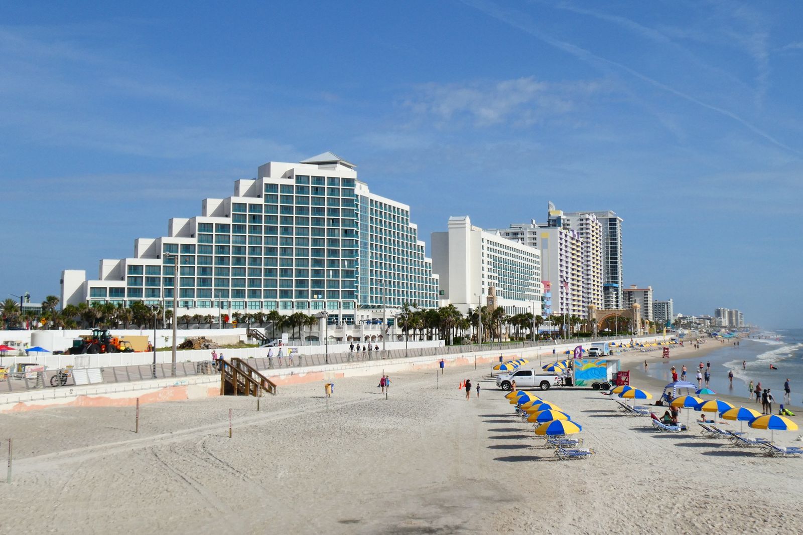 Daytona Beach seen from the Main Street Pier, with blue-and-yellow umbrellas on the sand and the Hilton Daytona Beach Oceanfront Resort behind