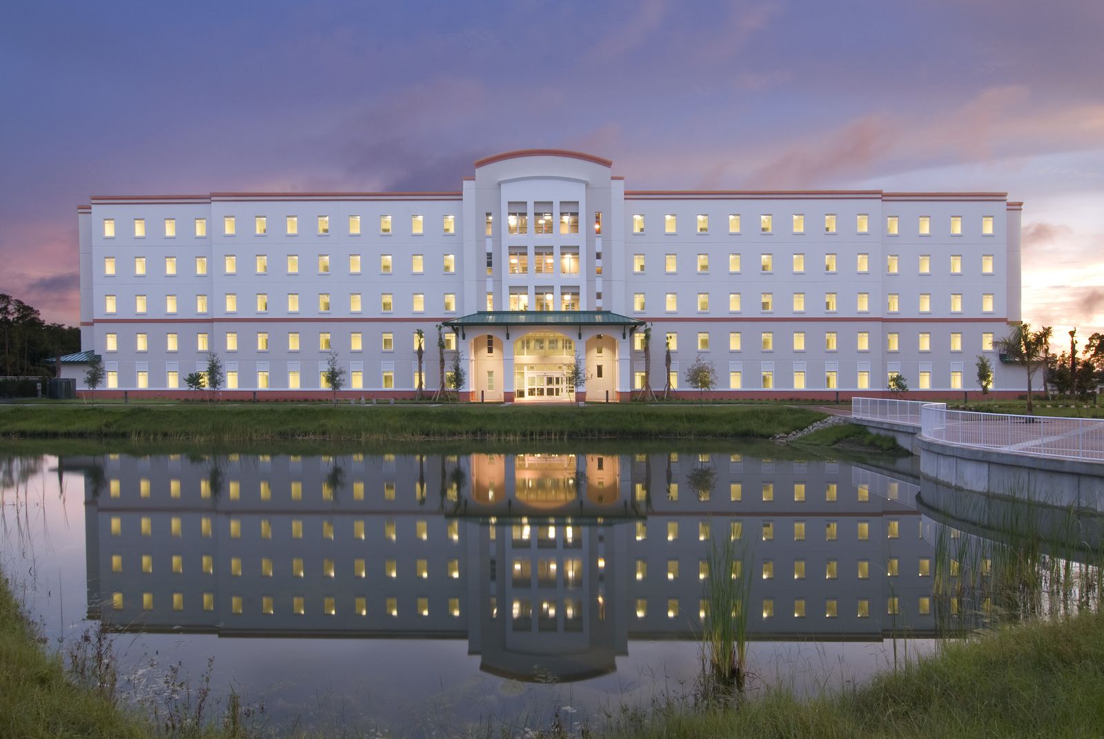FGCU campus building with its lights on at dusk, reflected in the lake in front under a pink sky