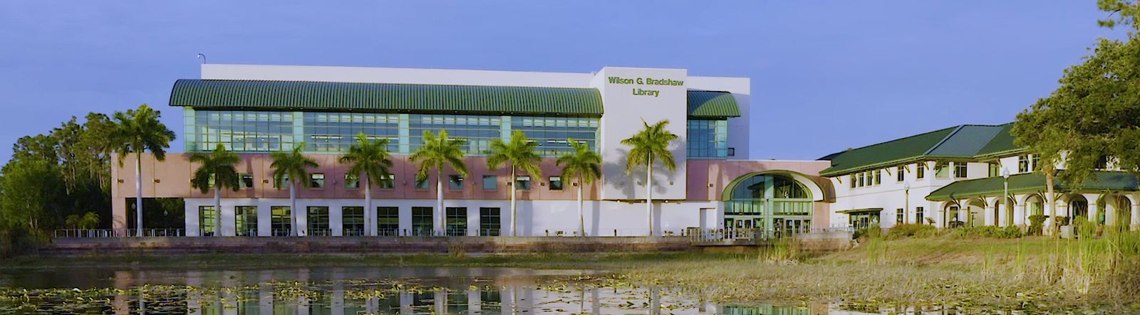 The green-roofed Wilson G. Bradshaw Library with palm trees, seen across the campus lake at Florida Gulf Coast University in Fort Myers on a blue-sky day
