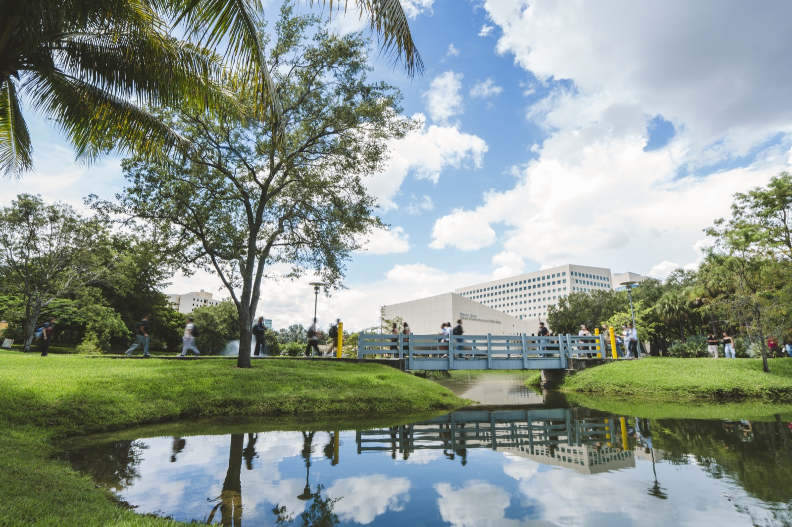 FIU campus lake with a wooden footbridge, trees and the Green Library behind, reflected in the water on a sunny day