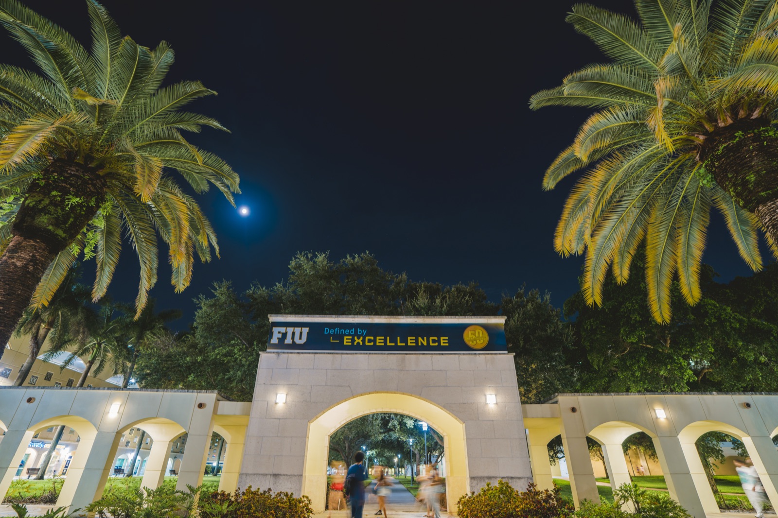 Entrance arch with the FIU Defined by Excellence sign lit at night, between palm trees under the moon