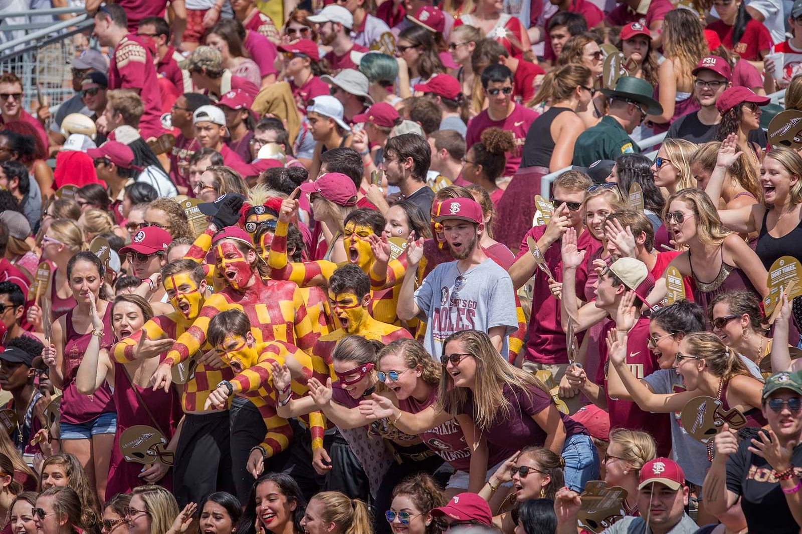 FSU students cheering in the student section of Doak Campbell Stadium at a football game, dressed in garnet and gold, some in body paint, on a sunny day in Tallahassee