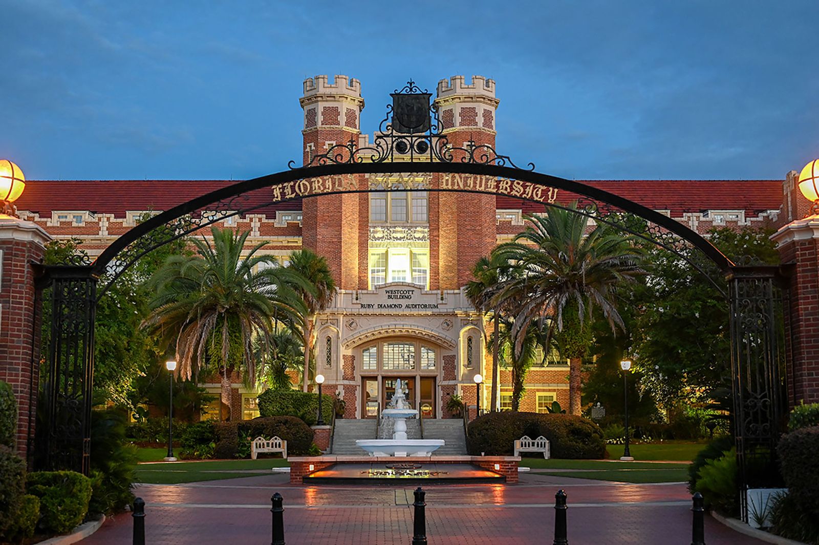 The Florida State University iron gateway lit at dusk, with palms, the fountain and the towers of the Westcott Building behind