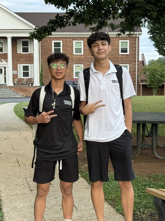 Dois alunos internos com mochilas e polo da escola em frente ao alojamento de tijolos