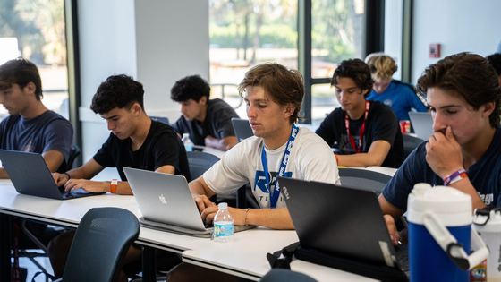 Meninos com camisetas da RPS trabalhando em laptops em uma sala de aula clara, com palmeiras do lado de fora