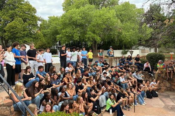 Alunos e professores sentados nos degraus do anfiteatro de pedra, sob álamos, aplaudindo em reunião da escola