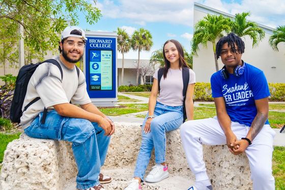 Três estudantes sentados em um muro de pedra coral diante do letreiro do Broward College no South Campus. com palmeiras e céu azul ao fundo