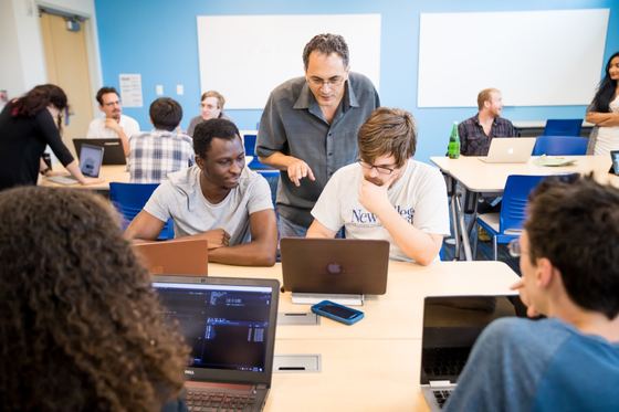 Professor orientando dois estudantes com laptops em uma sala de aula da New College of Florida. com outros alunos trabalhando em equipe ao redor