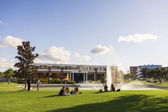 Estudantes sentados no gramado diante do Reflecting Pond. com a fonte e o Millican Hall ao fundo. no campus principal da UCF em Orlando