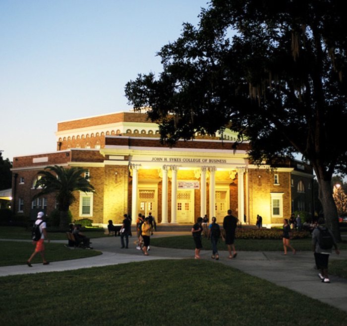 Fachada iluminada do John H. Sykes College of Business ao anoitecer, com estudantes no gramado em frente