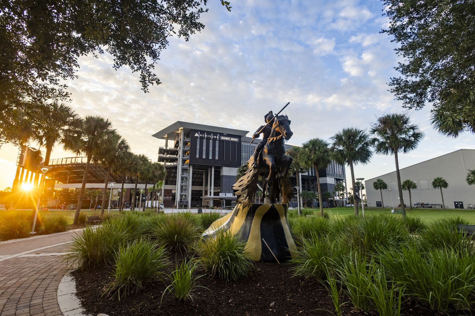 The Charging Knight statue at sunset, among palm trees, with Addition Financial Arena behind, at UCF's Knights Plaza