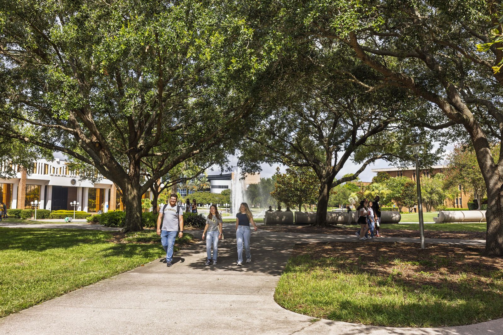 Students walking along a path under live oaks on UCF's main campus, with academic buildings behind