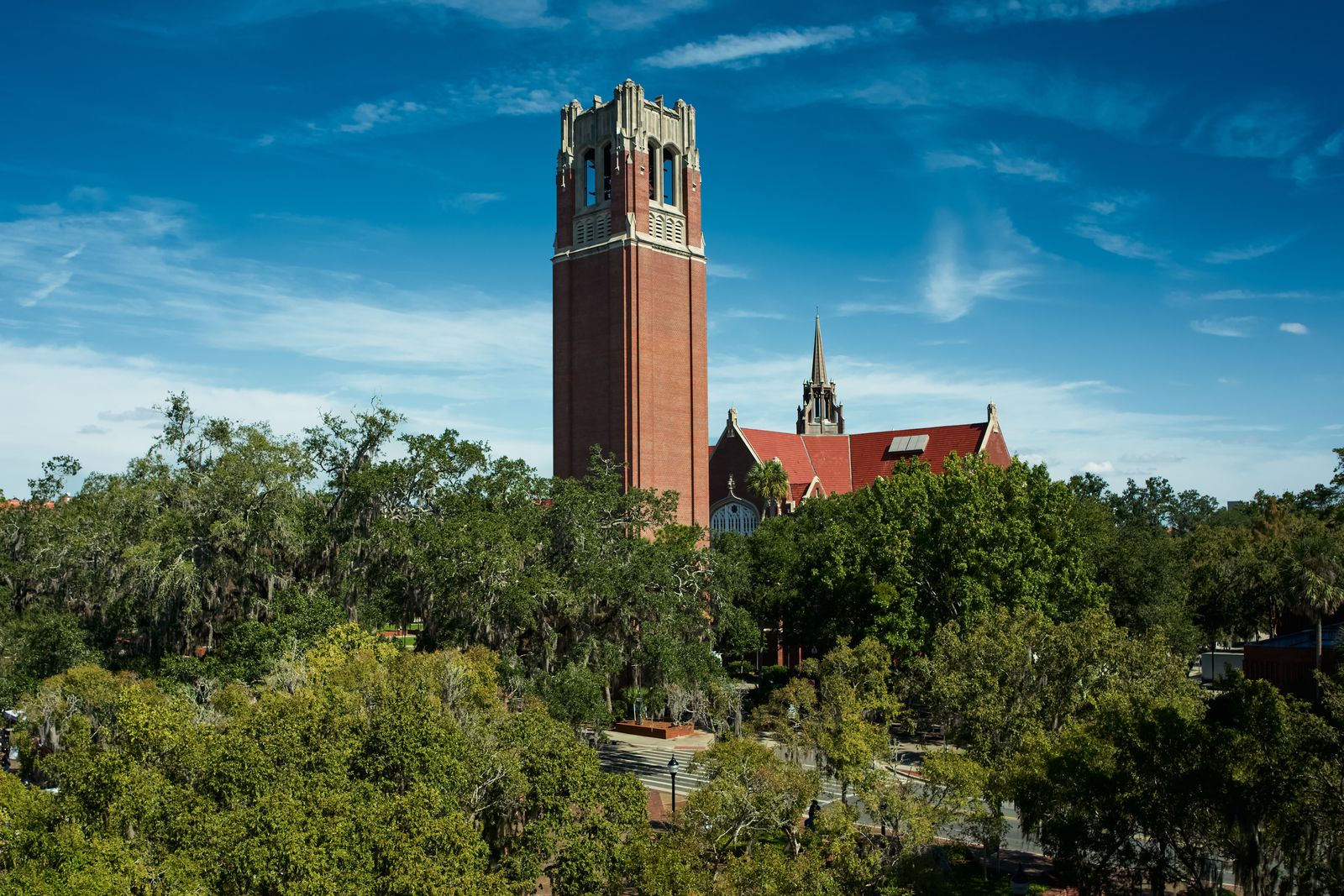 Century Tower, the University of Florida's red-brick carillon tower, rising above the campus oaks next to the University Auditorium under a blue sky in Gainesville