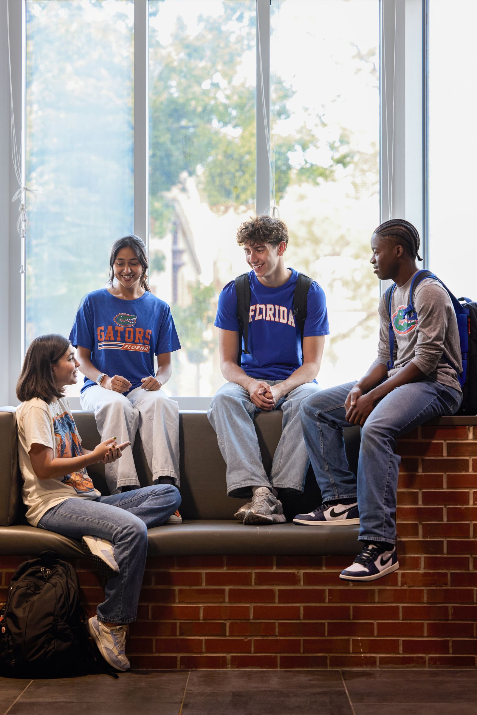 Four students in Florida and Gators T-shirts chatting on a brick window seat by a tall window in a common area on the University of Florida campus