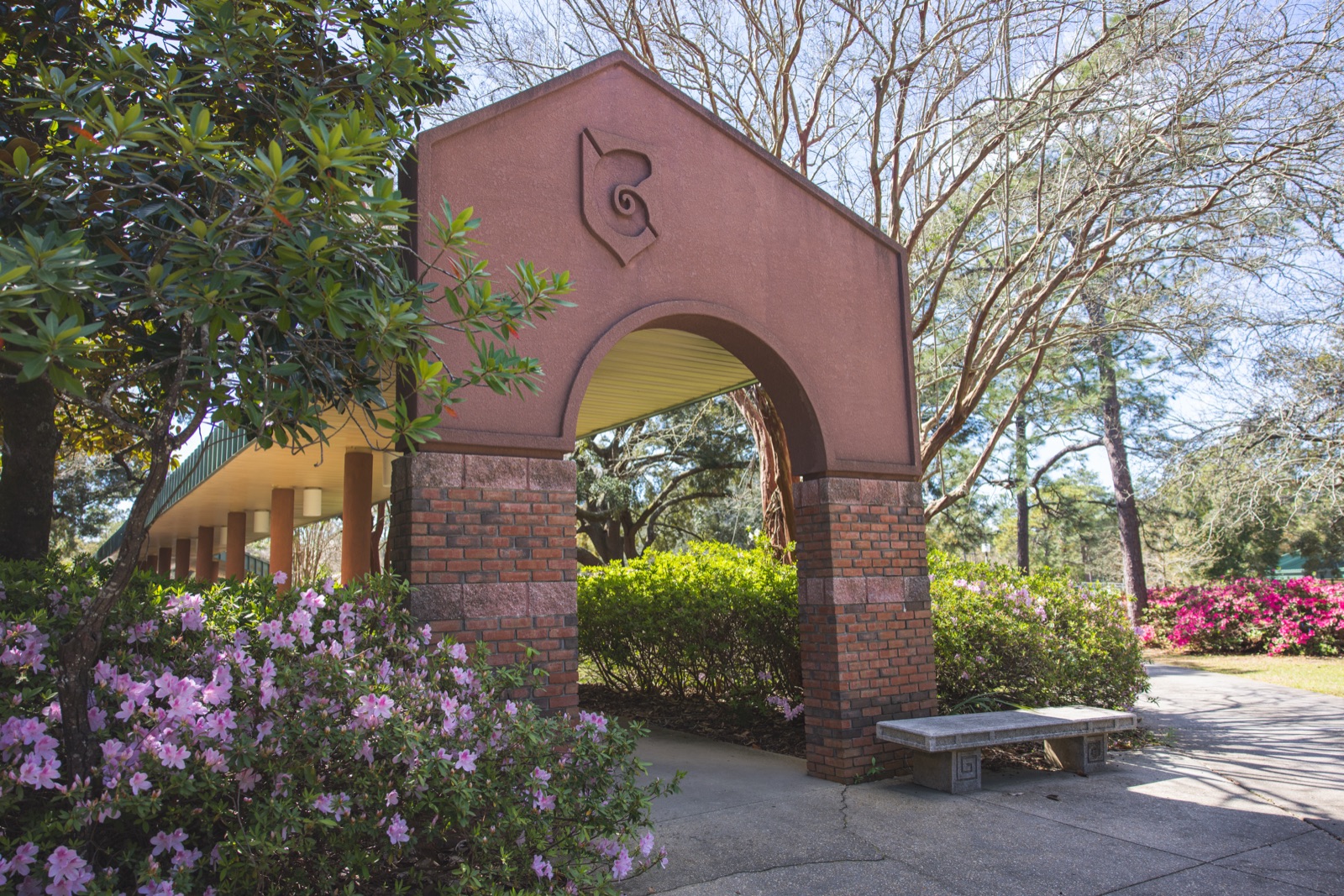 Brick arch bearing UWF's nautilus symbol among blooming azaleas and oaks on the Pensacola campus in spring