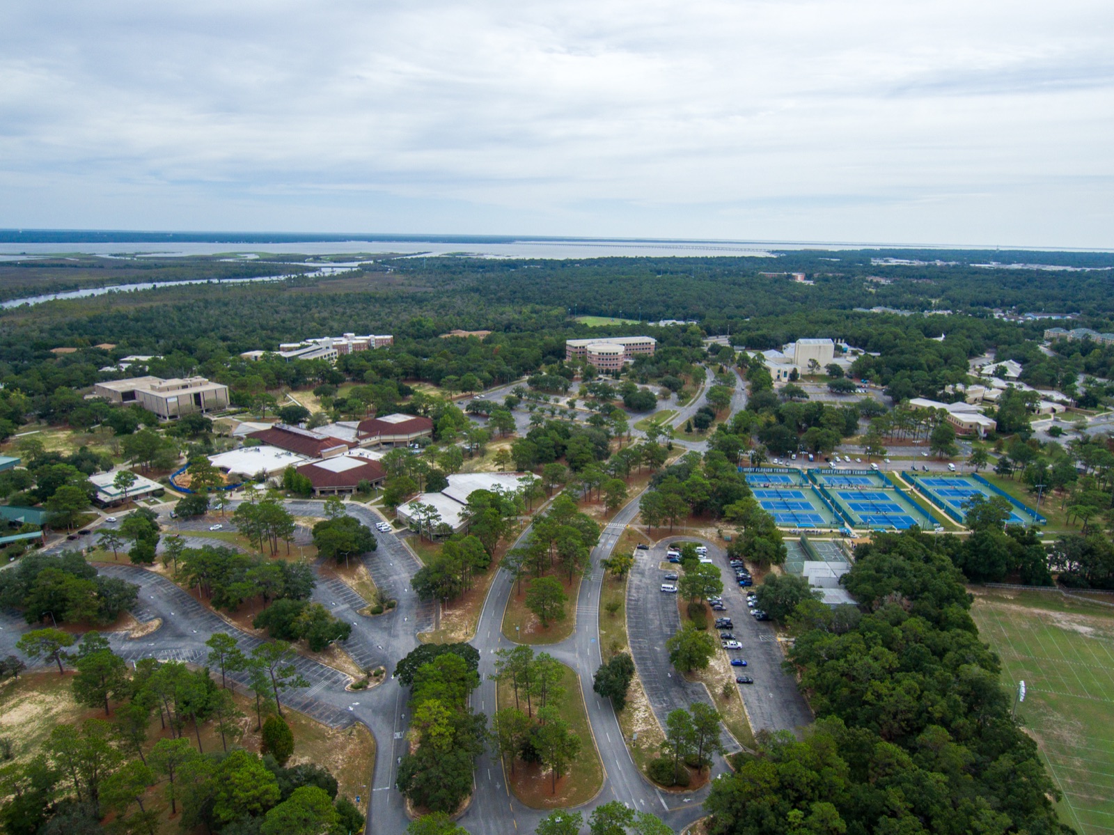 Aerial view of the University of West Florida campus in Pensacola, with the buildings among pine trees, the tennis courts and the bay in the background