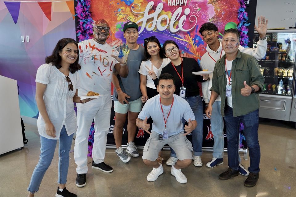 Estudantes e funcionários sorriem cobertos de pó colorido na celebração do Holi na Atlantis University, diante de um painel “Happy Holi”