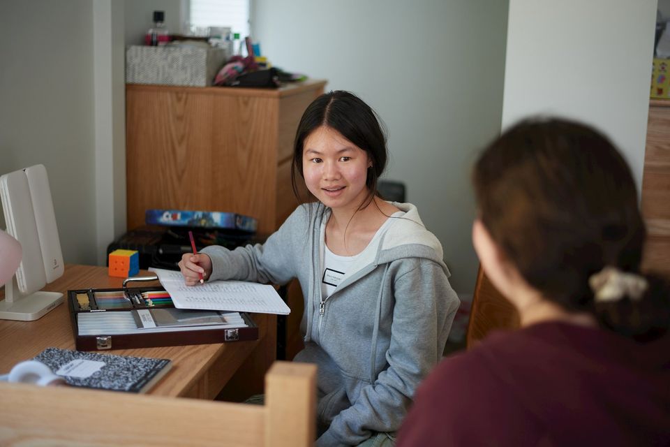Aluna estudando na escrivaninha do quarto do alojamento enquanto conversa com uma colega