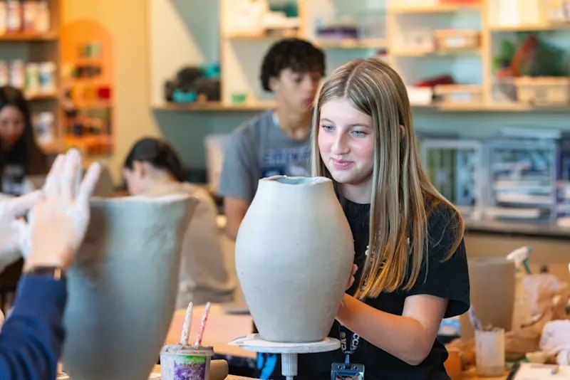 Menina modelando um vaso grande de argila em uma sala de cerâmica movimentada
