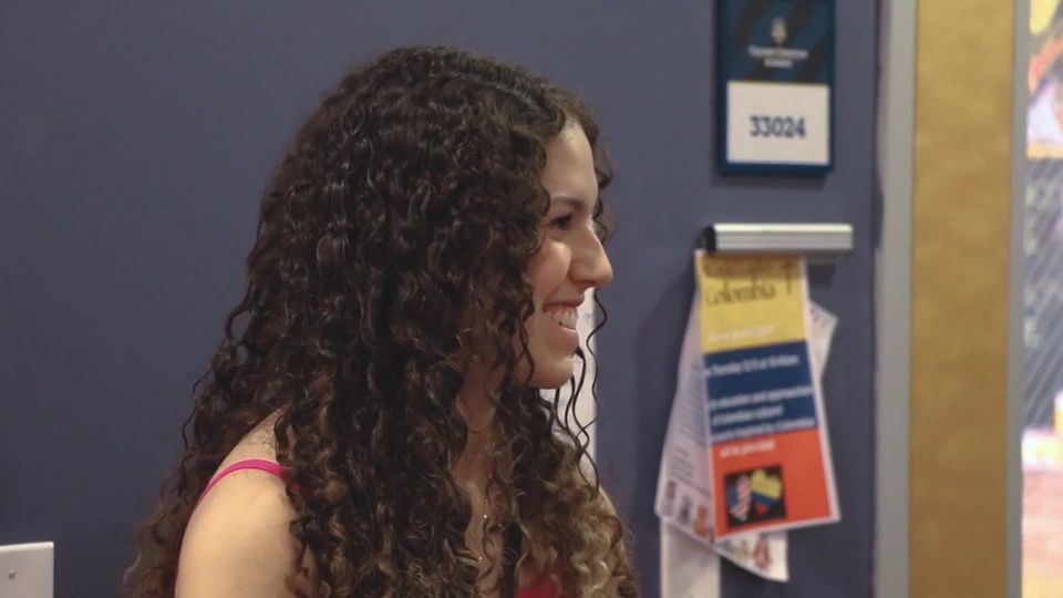 Sara, aluna internacional brasileira, sorrindo em um corredor da escola (quadro do vídeo de depoimento)