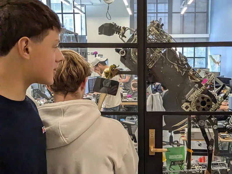 Dois alunos observando um braço robótico através da porta de vidro de um laboratório