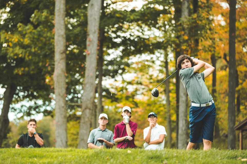 Golfista júnior de camiseta da GPA finalizando um drive enquanto o técnico e três colegas observam, com árvores de outono ao fundo