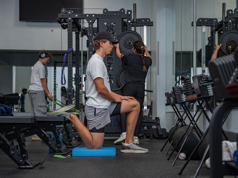 Três alunos treinando na academia de musculação (alongamento, elástico e barra)