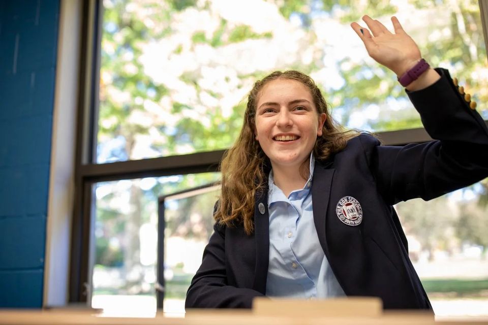 Aluna de blazer acenando sentada à mesa junto a uma janela