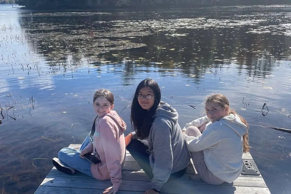 Três alunas sentadas em um píer de madeira à beira de um lago