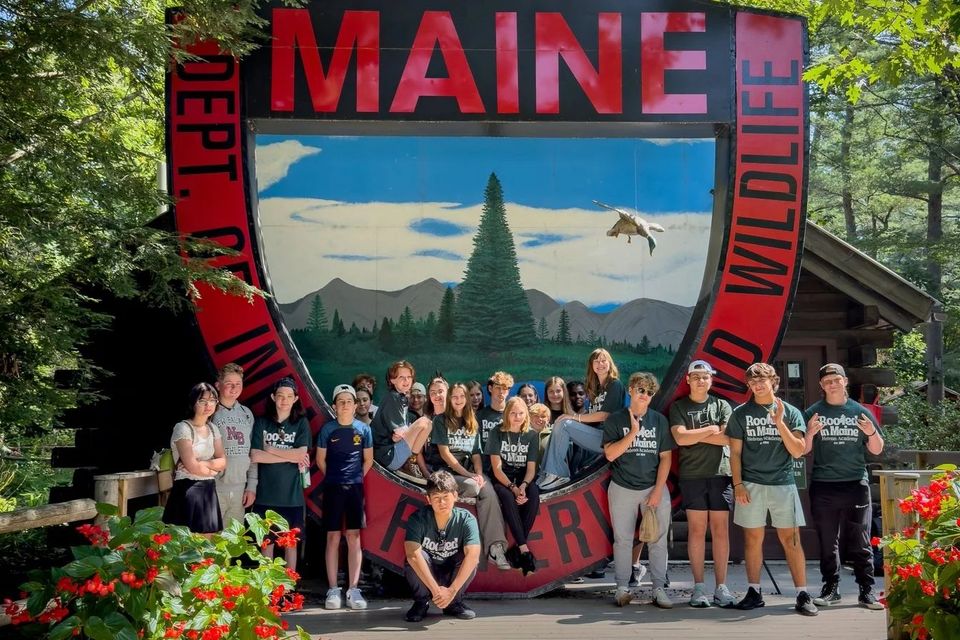 Grupo de alunos de camiseta verde diante de um painel do departamento de vida selvagem do Maine
