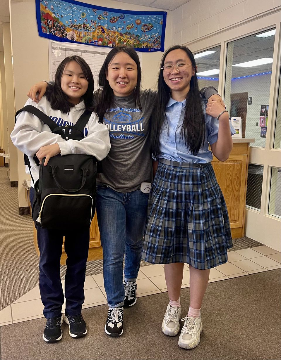 Três alunas abraçadas no corredor da escola, uma com camiseta de vôlei da Houghton Academy