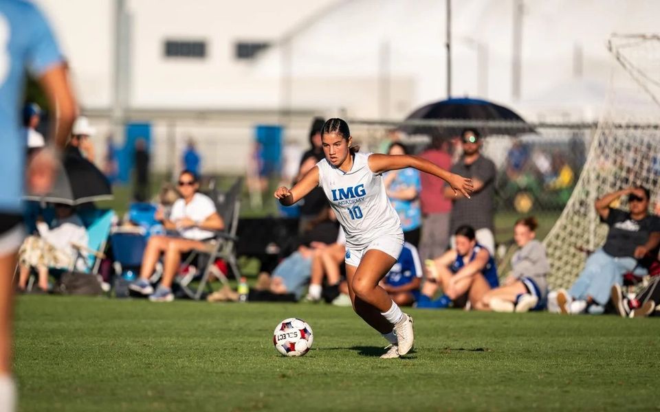 Jogadora de futebol com a camisa 10 branca da IMG Academy conduzindo a bola na IMGA Girls Cup, com público ao fundo