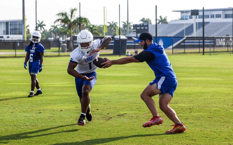 Técnico entregando a bola a um jogador de futebol americano da IMG durante o treino, com o estádio ao fundo
