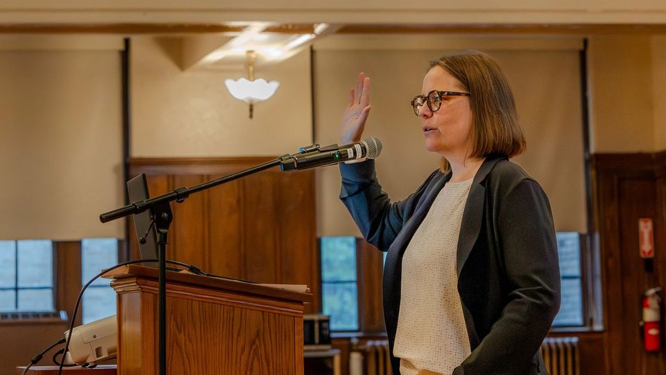Professora falando ao microfone em um púlpito de madeira no auditório
