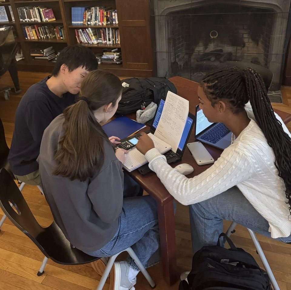 Três alunos estudando com laptops e cadernos diante de uma lareira na biblioteca
