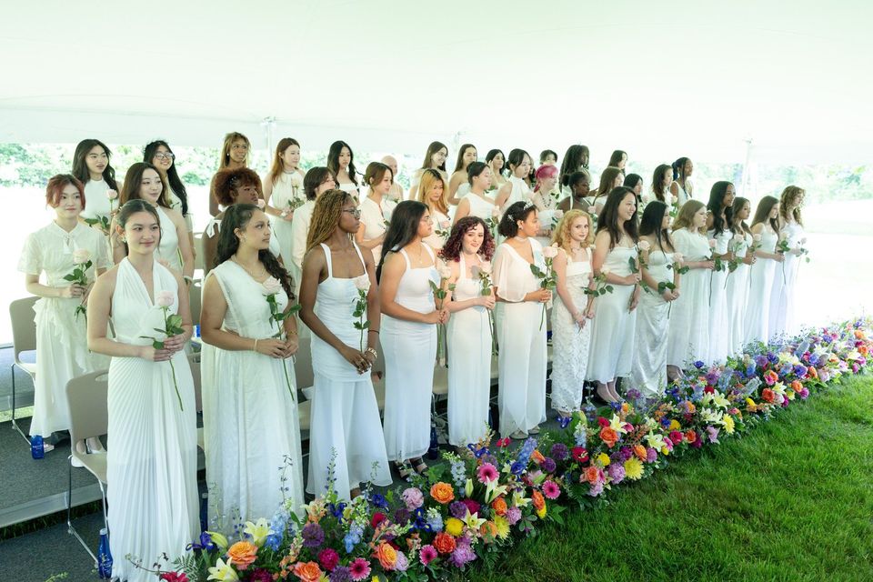 Formandas de vestido branco com rosas enfileiradas na cerimônia de formatura ao ar livre