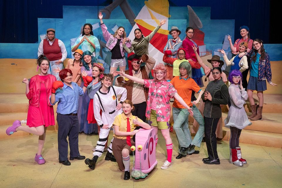 Elenco de alunas em um musical de inverno no palco, com cenário colorido