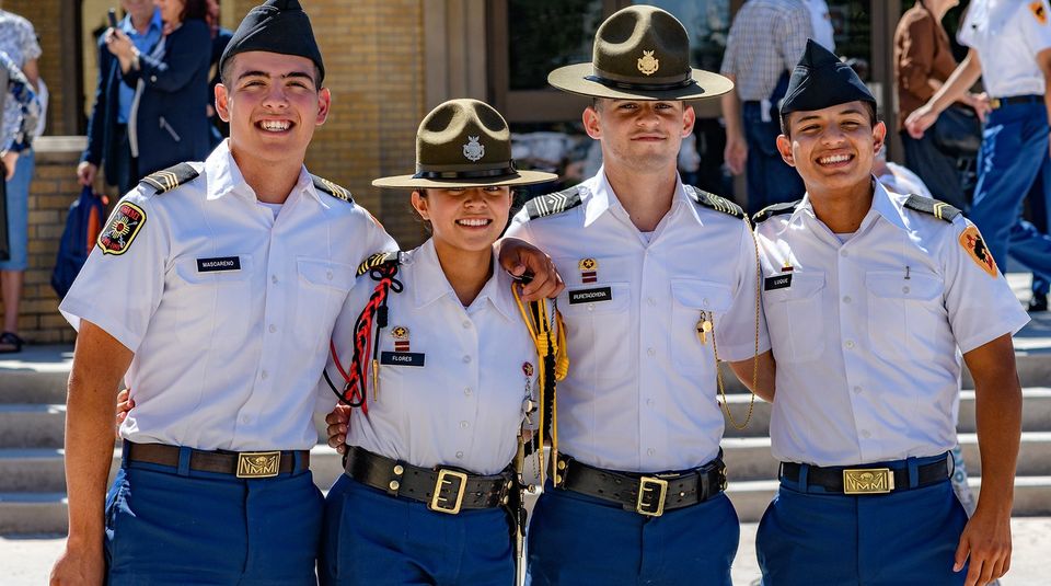Quatro cadetes sorrindo em uniforme branco de verão na escadaria de um prédio do campus