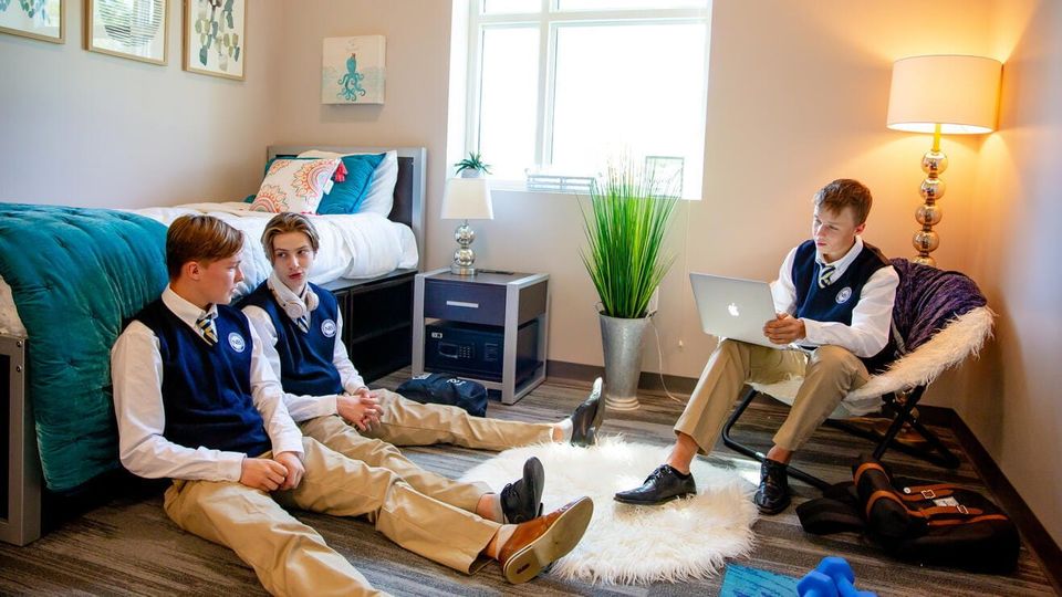 Três meninos de uniforme escolar em um quarto do dormitório, um deles no laptop