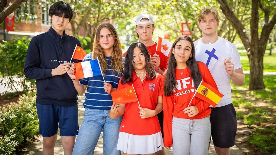 Seis alunos internacionais segurando bandeiras da França, China, Espanha e Finlândia sob árvores no campus