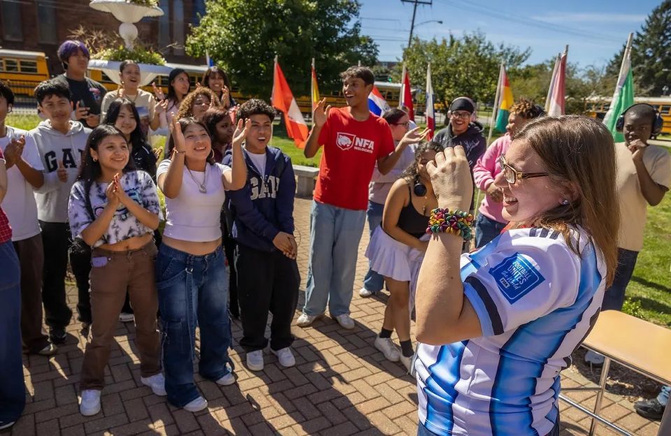 Alunos tirando uma selfie ao ar livre diante de bandeiras internacionais em um evento no campus
