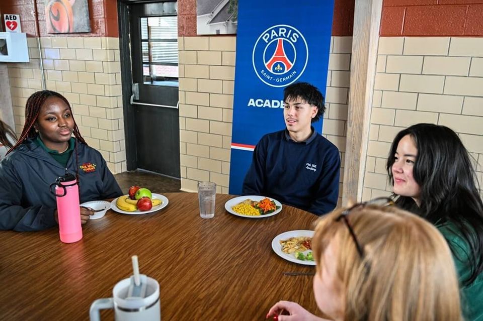 Alunos almoçando no refeitório ao lado de um banner da PSG Academy