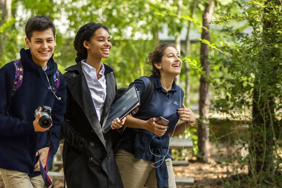 Três alunos de uniforme caminhando e sorrindo em uma trilha arborizada no campus