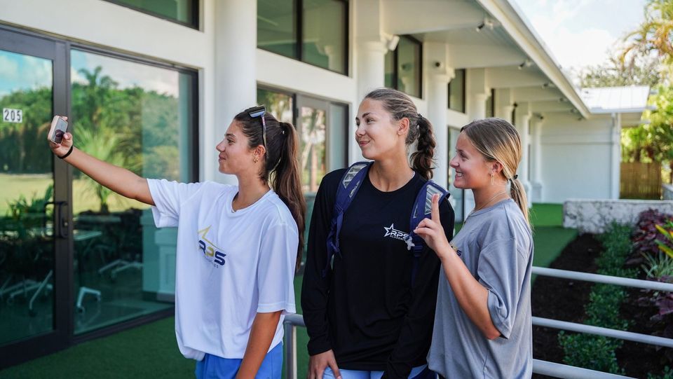 Três meninas com camisetas da RPS Academies tirando uma selfie em frente ao prédio acadêmico de colunas brancas